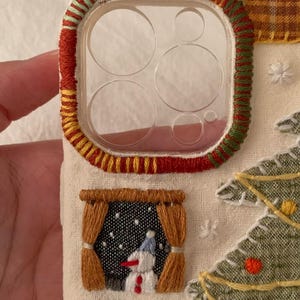 Op de afbeelding: Geborduurde telefoonhoes met een kerstthema. De hoes heeft een doorzichtige lensbeschermer met rode, gele en groene stiksels. Een klein raam toont een sneeuwpop en er is ook een kerstboom geborduurd.