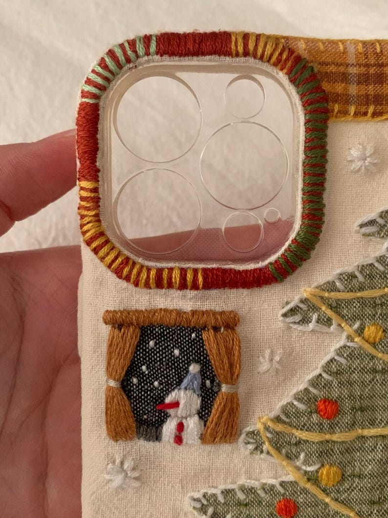 Op de afbeelding: Geborduurde telefoonhoes met een kerstthema. De hoes heeft een doorzichtige lensbeschermer met rode, gele en groene stiksels. Een klein raam toont een sneeuwpop en er is ook een kerstboom geborduurd.