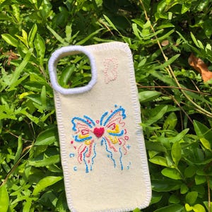 May include: A cream-colored fabric phone case featuring a hand-embroidered butterfly design in blue, yellow, and red thread. The center of the butterfly is a textured red heart. The edges and camera cutout are finished with white blanket stitching.