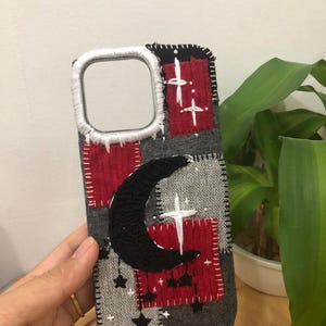 May include: A rectangular phone case featuring a patchwork design of red, grey, and black fabric squares. The case is decorated with hand-stitched embroidery of a large black crescent moon, white stars, and cross shapes, with white stitching around the camera.