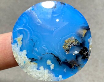 May include: A round, polished gemstone with a vibrant blue hue, reminiscent of a clear sky. The stone features swirling patterns of white, black, and gold inclusions, creating a unique and natural design. The gemstone is held by a finger.