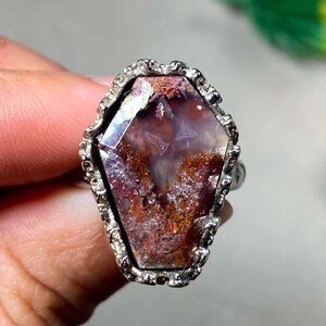 May include: A coffin-shaped ring featuring a faceted stone with deep red, brown, and translucent white inclusions. The stone is set within a textured, metallic silver-toned frame that follows the angular, six-sided outline of the gemstone.