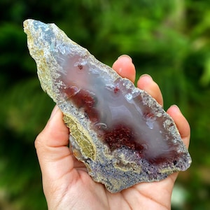 Könnte beinhalten: Eine Hand hält einen polierten Achatstein, der eine natürliche, unregelmäßige Form aufweist. Der Stein zeigt eine Mischung aus Farben, darunter durchscheinendes Grau, tiefes Rot und Anklänge von Gelb und Lila. Die Oberfläche des Achats ist glatt und poliert.