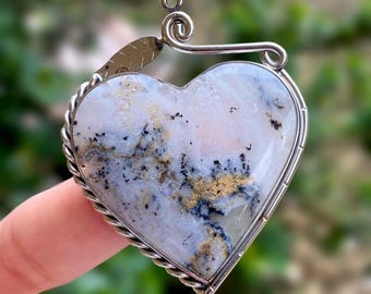 Puede incluir: Colgante en forma de corazón con marco y fianza plateados. El colgante presenta una piedra azul claro y blanca con inclusiones negras y doradas. El marco tiene un diseño de cuerda trenzada y una hoja decorativa.