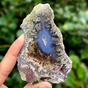 May include: A natural agate stone slice with a central blue cavity. The stone features a mix of colors, including blue, brown, green, and purple, with a textured, organic appearance. The agate is held against a blurred green background.
