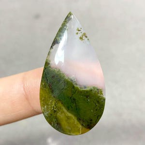 Può includere: Una gemma a forma di goccia lucidata. La pietra presenta una miscela di colori, tra cui trasparente, bianco, rosa e varie tonalità di verde. Le aree verdi assomigliano a muschio o inclusioni vegetali, creando un aspetto naturale.