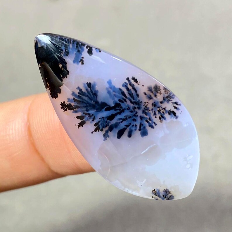 May include: A polished, elongated gemstone with a dendritic pattern. The stone features a base of pale lavender with intricate, fern-like inclusions in shades of deep blue and black. The overall shape is smooth and teardrop-like, ideal for jewelry.