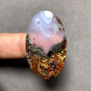 Peut inclure: Une pierre précieuse ovale avec un motif de paysage. La pierre présente un mélange de couleurs, notamment blanc, rose, vert, marron et or. Le motif évoque une scène naturelle miniature, avec un ciel nuageux et un terrain coloré.