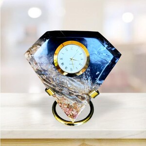 May include: A diamond-shaped desk clock with a gold-tone clock face and hands. The clock face has Roman numerals. The clock's body is a mix of blue, black, and brown tones. It has a gold-tone ring base.