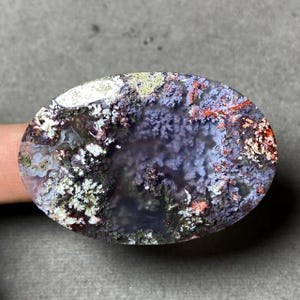 May include: An oval-shaped gemstone with a unique, natural pattern. The stone features a mix of colors, including white, purple, red, and green, creating a landscape-like appearance. The intricate details and color variations make it a striking piece.