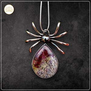 May include: A silver spider pendant necklace with a teardrop-shaped stone. The stone has red, purple, and gold colours. The spider's legs are curved, creating a unique design. The necklace is suspended from a silver chain.