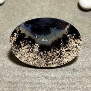 May include: Oval-shaped polished stone with a dark blue and black center, surrounded by a cream-colored, speckled border. The stone has a natural, organic appearance, with a smooth, glossy surface. The overall effect is a blend of dark and light tones.