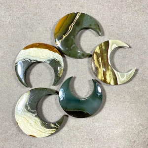 May include: Five polished crescent moon-shaped gemstones in shades of green, brown, and cream. The stones have unique patterns and textures, with variations in colour and veining. These decorative stones are ideal for jewellery making or display.