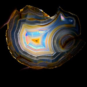 May include: A cross-section of a polished agate stone, showcasing concentric bands of color. The stone features a central, geometric pattern with hues of yellow, blue, and orange, surrounded by layers of white, gray, and brown. The background is black.