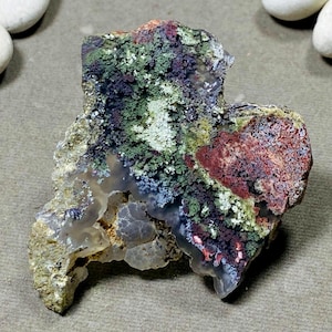 May include: A natural, irregularly shaped stone with a variety of colors. The stone features areas of green, red, and white, with a translucent, crystalline base. The overall texture is rough and organic, showcasing the stone's natural beauty.
