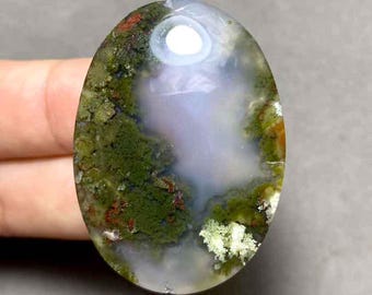 May include: An oval-shaped gemstone with a translucent, polished surface. The stone features a blend of green, white, red, and clear colors, resembling a landscape with mossy textures. The gemstone is held in a hand.