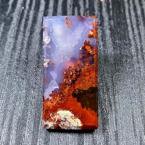 Puede incluir: Una gema rectangular con una superficie pulida. La piedra presenta una mezcla de colores, incluyendo rojo, naranja, blanco y morado. El diseño se asemeja a un paisaje con patrones orgánicos y ramificados. La gema está sobre un fondo oscuro.