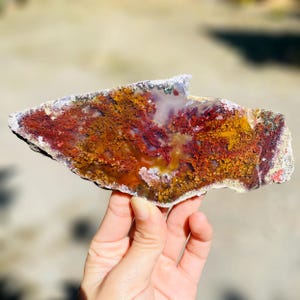 Könnte beinhalten: Eine Natursteinplatte mit einer lebendigen Farbgebung. Der Stein weist eine Mischung aus roten, gelben und orangen Farbtönen mit komplizierten Mustern auf, die einer Landschaft ähneln. Die Kanten sind rau und strukturiert, was zu seinem natürlichen Reiz beiträgt. Der Stein wird von einer Hand gehalten.