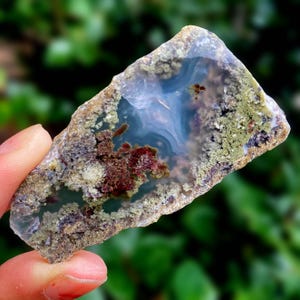May include: A polished agate stone slice with a translucent blue centre, surrounded by layers of brown, green, and red mineral deposits. The stone has a rough, natural edge and displays intricate patterns.