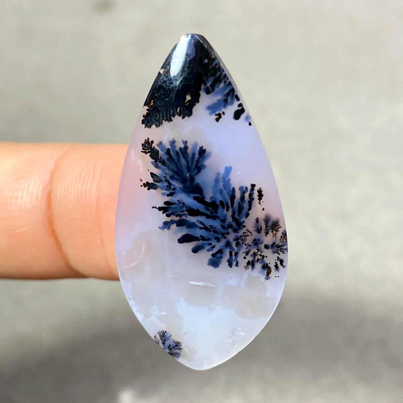 May include: A teardrop-shaped gemstone with a translucent white base, featuring intricate dendritic inclusions in shades of deep blue and black. The patterns resemble delicate, fern-like formations, creating a natural, artistic design.