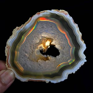 May include: A cross-section of a geode, displaying concentric bands of colour. The interior features a cavity with a dark opening and a central, glowing area. The outer edges show a mix of white, brown, and grey tones, with a rainbow-like iridescence.