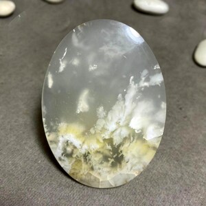 Pode incluir: Uma pedra preciosa ovalada e translúcida com uma aparência nublada e em camadas. A pedra apresenta inclusões brancas e amarelas, assemelhando-se a uma paisagem de nuvens. A superfície polida reflete a luz, destacando os padrões naturais.