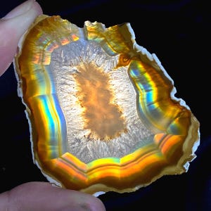 May include: A cross-section of a geode, showcasing a spectrum of colors. The outer edge is a warm brown, transitioning to bands of yellow, orange, and blue. The center features a crystalline structure with a light brown core. The geode is held by a finger.