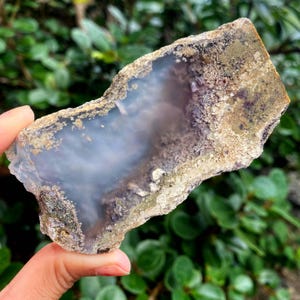 May include: A natural stone specimen with a light blue interior and a textured, multi-toned exterior. The stone features shades of beige, brown, and hints of purple. The edges are uneven, showcasing the raw, organic form of the mineral.