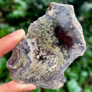 Op de afbeelding: Een natuurlijke steen met een onregelmatige vorm en een verscheidenheid aan kleuren. Het heeft een doorschijnend, lichtgrijs gebied, een dieprode sectie en gebieden van groen en wit, met een textuur, mosachtig uiterlijk. De steen wordt door een hand vastgehouden.