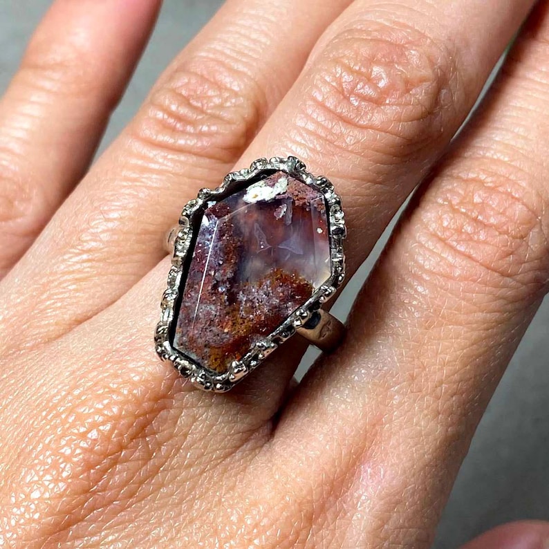 May include: A silver-toned ring featuring a large, coffin-shaped stone with a translucent, mottled surface displaying deep reddish-brown, white, and grey inclusions. The stone is set within a textured, scalloped metal bezel with a polished finish.