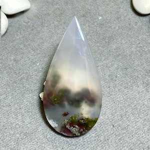 Può includere: Una gemma a forma di goccia con una base bianca lattea traslucida. La pietra presenta inclusioni interne di verde, rosso e marrone, che ricordano un paesaggio. La superficie lucida riflette la luce, evidenziando i motivi naturali. Per gioielli.