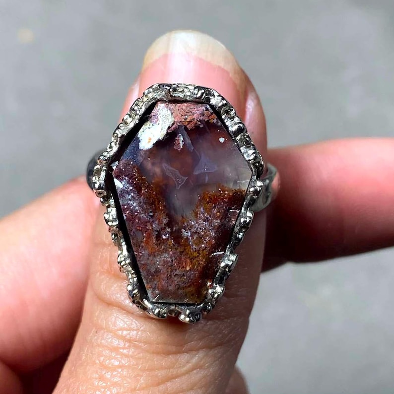 May include: A silver ring featuring a coffin-shaped stone with a textured, metallic setting. The stone displays a mix of deep reddish-brown, translucent white, and earthy tones with a crystalline, speckled surface texture throughout the center.