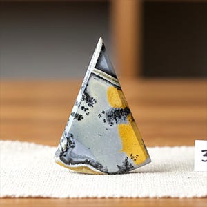 Puede incluir: Una piedra triangular pulida con un patrón único. La piedra presenta una mezcla de colores grises, amarillos y negros. La piedra está de pie sobre una superficie blanca. La forma general es una pirámide.