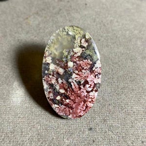 May include: An oval gemstone with a distinctive pattern. The stone displays a blend of red, green, and white hues, forming a natural, organic design. The gemstone is polished and smooth, with a translucent quality.