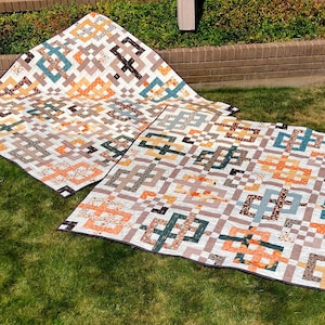 May include: Two rectangular quilts featuring a repeating geometric pattern of interlocking squares. The fabric displays a white background with printed motifs in shades of orange, teal, brown, and grey, finished with a dark grey binding edge.