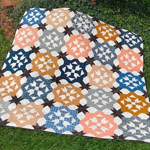 May include: A colorful quilt with a geometric starburst pattern. The quilt features a variety of colors, including blue, peach, gold, and gray, set against a dark brown background. The quilt is made of fabric and is likely handmade.
