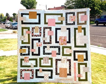 May include: A square quilt featuring a white background with a geometric pattern of interlocking rectangular frames in olive green and black. The design incorporates various fabric squares and rectangles in shades of pink, orange, and brown.