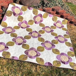 May include: A handmade quilt featuring a repeating floral pattern. The quilt showcases large, stylized flowers in shades of purple and cream, with olive green leaves. The background is white with a subtle, quilted texture.