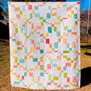 May include: A white quilt with a patchwork design featuring squares of various colors and patterns, including pink, yellow, green, and blue. The quilt is quilted with a swirl pattern.