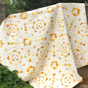 May include: A cream-colored quilt with a geometric pattern in shades of yellow and orange. The quilt features repeating square and star-like designs, with a quilted texture. The edges are finished with a yellow border.