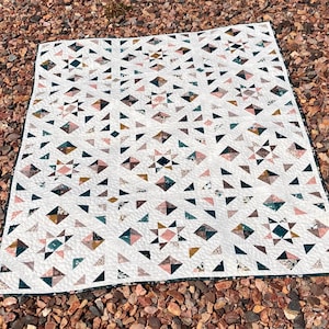 May include: A rectangular quilt featuring a repeating star and geometric pattern. The design consists of white fabric blocks interspersed with printed fabric in shades of dusty pink, dark teal, mustard yellow, and floral patterns, finished with a dark border.