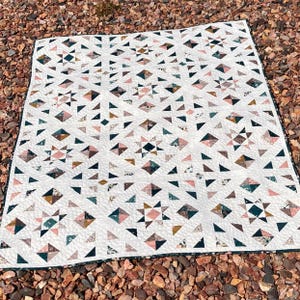 May include: A rectangular quilt featuring a repeating star and geometric pattern. The design consists of white fabric blocks interspersed with printed fabric in shades of dusty pink, dark teal, mustard yellow, and floral patterns, finished with a dark border.