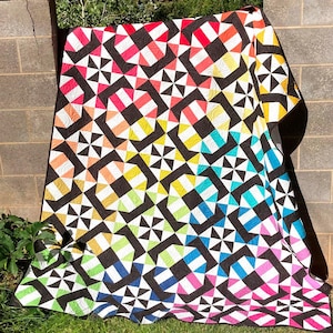 May include: A colorful quilt with a geometric pattern of black, white, pink, red, orange, yellow, green, blue, and turquoise. The quilt is made up of squares and triangles, and the colors are arranged in a gradient pattern.