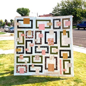 May include: A square quilt featuring a white background with a geometric pattern of interlocking rectangular frames in olive green and black. The design incorporates various fabric squares and rectangles in shades of pink, orange, and brown.
