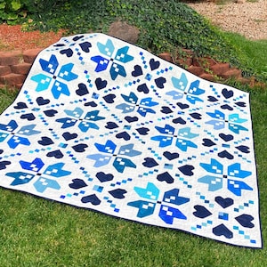 Pode incluir: Um edredom branco com um padrão de flocos de neve e corações azuis e azul marinho. O edredom, feito de tecido, tem uma borda azul escuro. É exibido na grama verde.