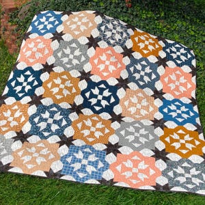 May include: A colorful quilt with a geometric starburst pattern. The quilt features a variety of colors, including blue, peach, gold, and gray, set against a dark brown background. The quilt is made of fabric and is likely handmade.