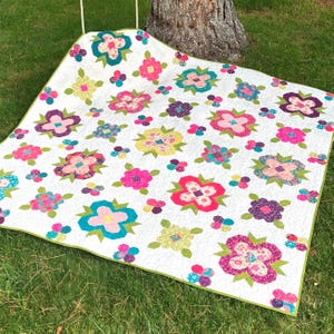 Puede incluir: Un edredón blanco con un estampado floral multicolor, que incluye rosa, azul, amarillo y morado. El edredón tiene un borde verde y está colocado sobre hierba verde. Las flores están dispuestas en un patrón repetitivo.