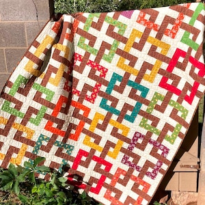 Op de afbeelding: Een kleurrijk quilt met een geometrisch patroon van vierkanten en rechthoeken in tinten bruin, oranje, geel, groen en rood. De quilt is gemaakt met verschillende stoffen, waaronder bloemenprints en effen kleuren.