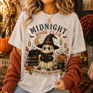May include: A light gray t-shirt featuring a graphic of a ghost wearing a witch hat and glasses, reading a book titled Potions & Spells. The design includes a black cat, pumpkins, candles, and the text Midnight Book Club and Reading Spells Since Forever.