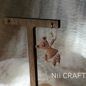 May include: A handmade brown crochet teddy bear earring with white accents, hanging from a gold-colored hook. The earring is displayed on a wooden earring stand. The text "Nii CRAFT" is visible.